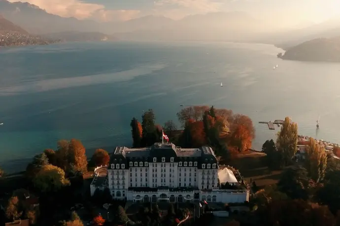 Vue aérienne d'un bâtiment historique au bord d'un lac entouré de montagnes, mettant en avant l'usage des drones pour promouvoir le patrimoine naturel et culturel.