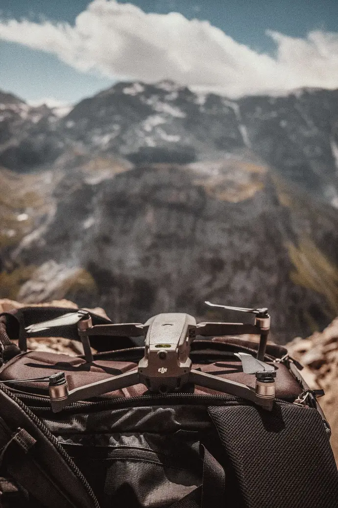 Drone posé sur un sac à dos, avec un paysage montagneux flou en arrière-plan sous un ciel partiellement nuageux